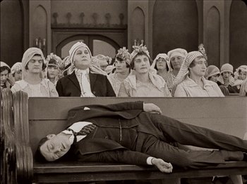 Movie still from “Seven Chances” (1925), directed by Buster Keaton – A man is lying on a bench in front of a group of people; Wide shot, High angle