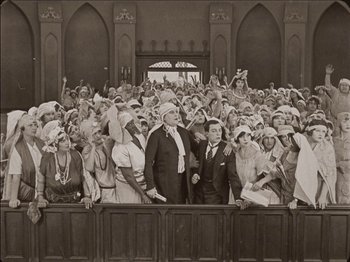 Movie still from “Seven Chances” (1925), directed by Buster Keaton – An old photo of a group of people standing in front of a building; Wide shot, High angle