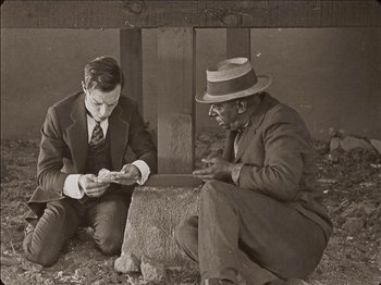 Movie still from “Seven Chances” (1925), directed by Buster Keaton – Two men sitting on the ground looking at papers; Medium shot, High angle