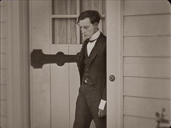 Movie still from “Seven Chances” (1925), directed by Buster Keaton – A man in a suit standing in front of an open door; Medium shot, High angle