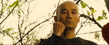 An older man with a bald head is standing in front of a tree