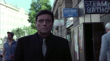 Movie still from “Sexy Beast” (2000), directed by Jonathan Glazer – A man wearing a suit and tie standing in front of a building; Close Up shot, Low angle