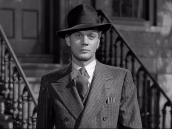 Movie still from “Shadow of a Doubt” (1943), directed by Alfred Hitchcock – A man in a suit and hat standing in front of stairs; Close Up shot, Low angle