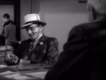 Movie still from “Shadow of a Doubt” (1943), directed by Alfred Hitchcock – A man in a suit and a hat sitting at a table; Medium shot, Over the shoulder angle