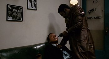 Movie still from “Shaft” (1971), directed by Gordon Parks – Two men in a room one is holding a gun and the other is sitting on a couch; Medium shot, Over the shoulder angle