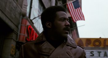 Movie still from “Shaft” (1971), directed by Gordon Parks – A man in a brown jacket standing in front of american flags; Close Up shot, Low angle