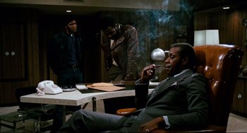 Movie still from “Shaft” (1971), directed by Gordon Parks – A man sitting in a chair smoking a cigarette while two other men watch; Medium shot, Low angle