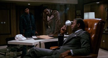 Movie still from “Shaft” (1971), directed by Gordon Parks – A man sitting in a chair holding a cigarette; Wide shot, Over the shoulder angle