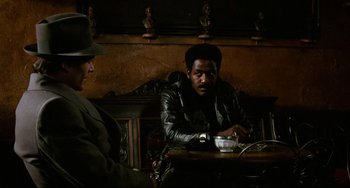 Movie still from “Shaft” (1971), directed by Gordon Parks – A man sitting at a table with a man in a hat; Medium shot, Over the shoulder angle