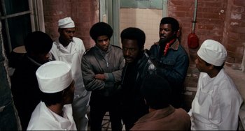 Movie still from “Shaft” (1971), directed by Gordon Parks – A group of men standing next to each other in front of a brick wall; Medium shot, High angle