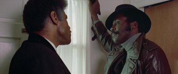 Movie still from “Shaft's Big Score!” (1972), directed by Gordon Parks – A man in a hat is holding a knife up to a man in a suit; Close Up shot, Over the shoulder angle