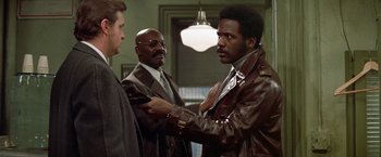 Movie still from “Shaft's Big Score!” (1972), directed by Gordon Parks – A man in a brown leather jacket holding another man in a suit; Medium shot, Over the shoulder angle