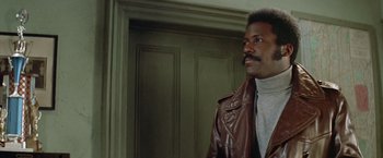Movie still from “Shaft's Big Score!” (1972), directed by Gordon Parks – A man in a brown leather jacket standing next to a door; Close Up shot, Low angle