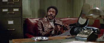 Movie still from “Shaft's Big Score!” (1972), directed by Gordon Parks – A man sitting on top of a red couch holding a remote control; Medium shot, Low angle