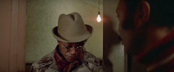 Movie still from “Shaft's Big Score!” (1972), directed by Gordon Parks – A man wearing a hat and glasses in a dimly - lit room; Close Up shot, Low angle
