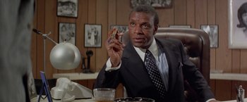 Movie still from “Shaft's Big Score!” (1972), directed by Gordon Parks – A man in a suit and tie sitting at a table with a cigarette in his hand; Medium shot, Low angle