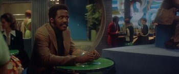 Movie still from “Shaft's Big Score!” (1972), directed by Gordon Parks – A man sitting at a table with a glass of water; Medium shot, Over the shoulder angle