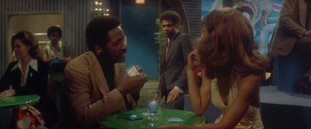 Movie still from “Shaft's Big Score!” (1972), directed by Gordon Parks – A man and a woman sitting at a table with two other people; Medium shot, Over the shoulder angle