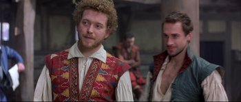 Movie still from “Shakespeare in Love” (1998), directed by John Madden – Two men in medieval garb are sitting and standing together; Medium shot, Over the shoulder angle