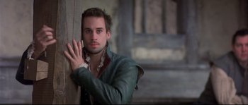 Movie still from “Shakespeare in Love” (1998), directed by John Madden – A man leaning against a wooden post in front of a building; Close Up shot, Over the shoulder angle