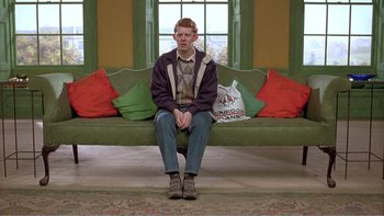 Movie still from “Shallow Grave” (1994), directed by Danny Boyle – A man sitting on top of a green couch in front of a window; Wide shot, High angle