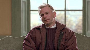 Movie still from “Shallow Grave” (1994), directed by Danny Boyle – A person sitting on a couch wearing a jacket; Close Up shot, Over the shoulder angle