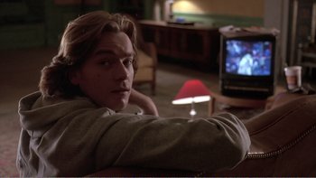 Movie still from “Shallow Grave” (1994), directed by Danny Boyle – A man sitting in front of a tv in a living room; Medium shot, Over the shoulder angle