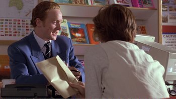 Movie still from “Shallow Grave” (1994), directed by Danny Boyle – A man sitting at a table with a woman holding papers; Medium shot, Over the shoulder angle