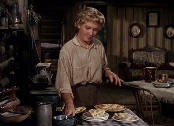 Movie still from “Shane” (1953), directed by George Stevens – A woman standing at a table with plates of food on top of it; Medium shot, Over the shoulder angle