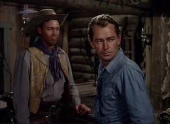 Movie still from “Shane” (1953), directed by George Stevens – A man in a cowboy hat standing next to another man in a blue shirt; Medium shot, Over the shoulder angle
