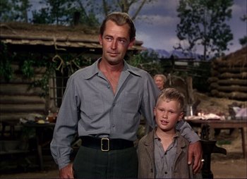 Movie still from “Shane” (1953), directed by George Stevens – A man standing next to a young boy in front of a log cabin; Medium shot, Low angle