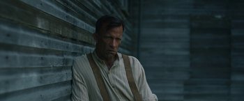 Movie still from “1922” (2017), directed by Zak Hilditch – A man in striped shirt and suspenders leaning against a wall; Medium shot, Low angle