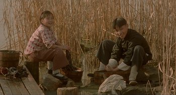 Movie still from “Shanghai Triad” (1995), directed by Yimou Zhang – Two people sitting on the ground near a body of water; Wide shot, High angle