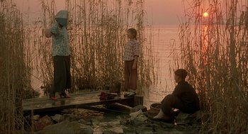 Movie still from “Shanghai Triad” (1995), directed by Yimou Zhang – Three people sitting on a dock near a body of water; Wide shot, Low angle