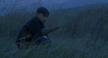 Movie still from “Shanghai Triad” (1995), directed by Yimou Zhang – A man sitting in the grass holding a stick; Medium shot, High angle