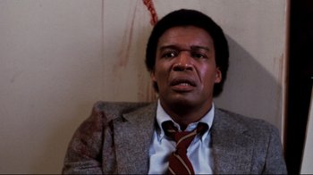 A man wearing a suit and tie with blood on the wall behind him