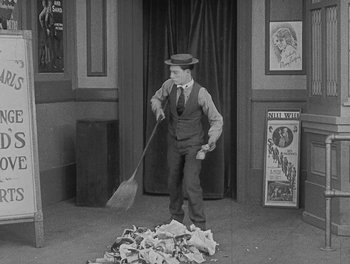 Movie still from “Sherlock Jr.” (1924), directed by Buster Keaton – A man in a top hat and vest holding a broom in front of a pile of papers; Wide shot, High angle