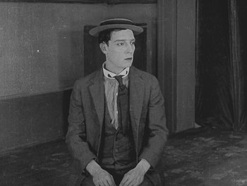 Movie still from “Sherlock Jr.” (1924), directed by Buster Keaton – A man wearing a suit and a hat sitting in a room; Medium shot, High angle