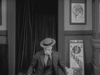 Movie still from “Sherlock Jr.” (1924), directed by Buster Keaton – A man in a suit and hat standing in front of a poster; Medium shot, High angle