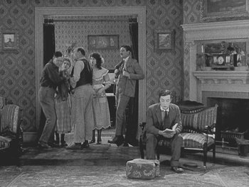 Movie still from “Sherlock Jr.” (1924), directed by Buster Keaton – A man sitting in a chair in front of a group of people in a living room; Wide shot, High angle