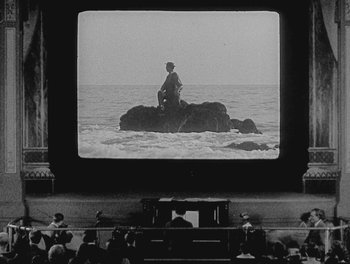 Movie still from “Sherlock Jr.” (1924), directed by Buster Keaton – People are sitting in front of a large screen television; Extreme Wide shot, Low angle