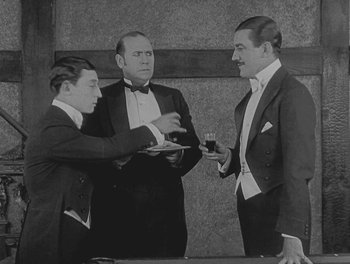 Movie still from “Sherlock Jr.” (1924), directed by Buster Keaton – Three men in tuxedos are standing around a table; Medium shot, Low angle