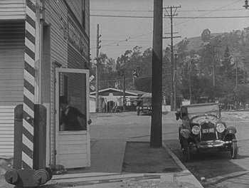 Movie still from “Sherlock Jr.” (1924), directed by Buster Keaton – An old photo of a street with a car parked on the side of the road; Extreme Wide shot, High angle