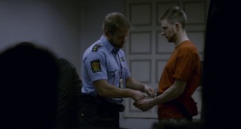 Movie still from “22 July” (2018), directed by Paul Greengrass – A police officer holding a man's hand while he is handcuffed; Medium shot, Over the shoulder angle