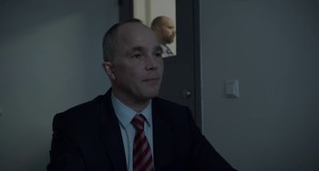 Movie still from “22 July” (2018), directed by Paul Greengrass – A man in a suit and tie sitting in front of a door; Medium shot, Over the shoulder angle