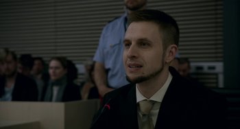 Movie still from “22 July” (2018), directed by Paul Greengrass – A man in a suit and tie is speaking at a meeting; Close Up shot, Over the shoulder angle