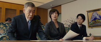 Movie still from “Shin Godzilla” (2016), directed by Shinji Higuchi – A woman sitting at a table in front of two men; Medium shot, Low angle