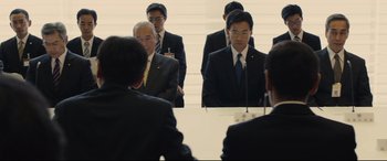 Movie still from “Shin Godzilla” (2016), directed by Shinji Higuchi – A group of men in suits standing in front of a mirror; Medium shot, Over the shoulder angle
