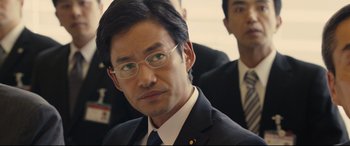Movie still from “Shin Godzilla” (2016), directed by Shinji Higuchi – A person wearing a suit and glasses; Close Up shot, Over the shoulder angle