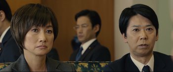 Movie still from “Shin Godzilla” (2016), directed by Shinji Higuchi – A man and a woman in a suit and tie in front of another man in a suit and tie; Close Up shot, Over the shoulder angle